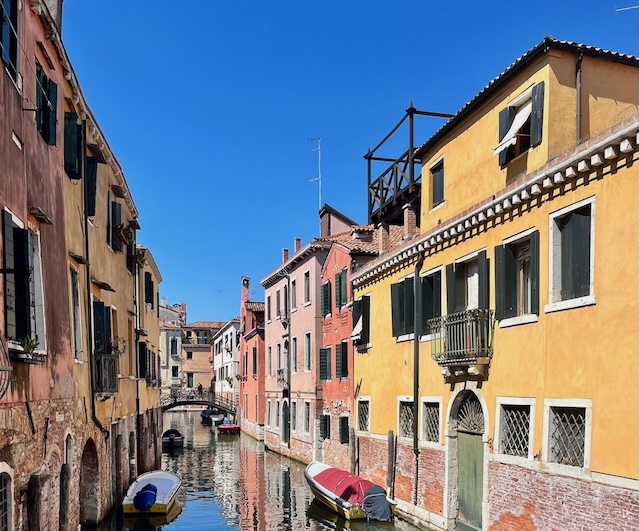 Picturesque canal in Venice, Italy, lined with colorful historic buildings and small boats moored along the waterway under a bright blue sky.