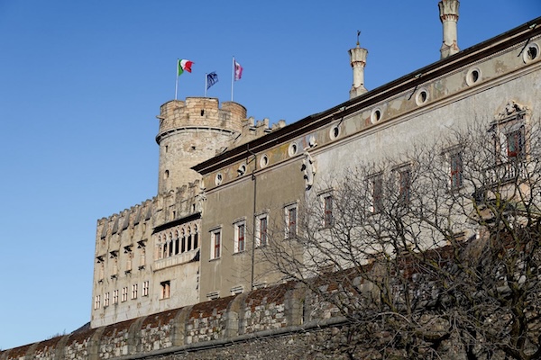 Historic Buonconsiglio Castle in Trento, Italy with Italian and regional flags flying above medieval stone towers under a clear blue sky