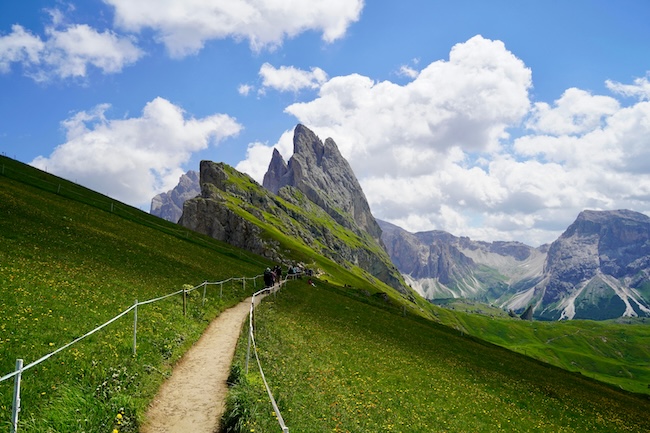 Hikers walking along a scenic trail toward the dramatic jagged peaks of Seceda in the Dolomites, surrounded by alpine meadows and mountain vistas