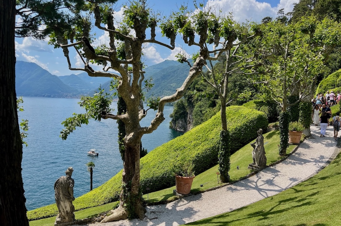 Scenic garden path with statues and trimmed trees at Villa del Balbianello on Lake Como, overlooking the lake and surrounding mountains.