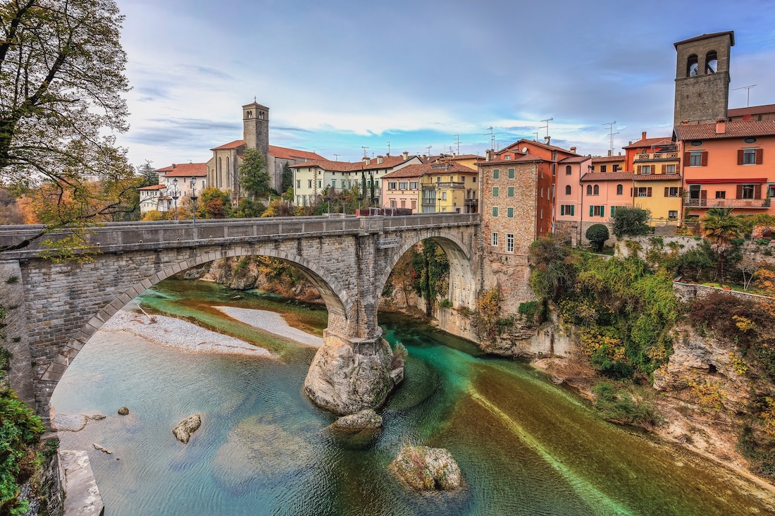 Historic stone Devil’s Bridge in Cividale del Friuli crossing the clear Natisone River with medieval buildings behind it.