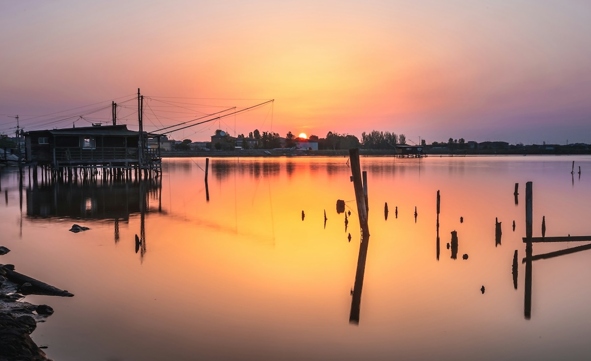 Traditional fishing huts and wooden posts reflected in the Po Delta wetlands at sunset, with warm orange and pink light over the lagoon