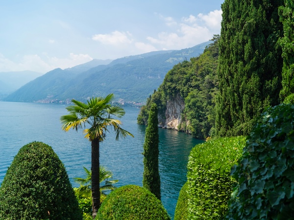 Lake Como’s shoreline with manicured gardens, cypress trees and mountain views on a sunny day in northern Italy.
